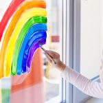 Kids at home. A child girl paints a rainbow on a window during the quarantine for the coronavirus pandemic. Social flash mob in support of society. Let’s all be well. Stay at home